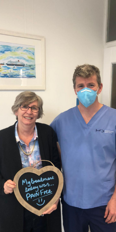 Patient with Dr Robin Hanna at Hampshire Dentists, holding a sign saying "My treatment today was pain free" emphasising pain-free dental care.