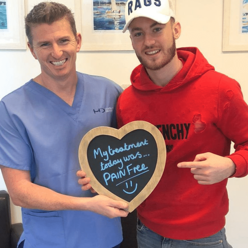 Dr Robin Hanna, Implantologist - Hampshire Dentists Pain Free Dentist in Southampton Patient with Dr Robin Hanna at Hampshire Dentists, holding a sign saying "My treatment today was pain free" emphasising pain-free dental care.