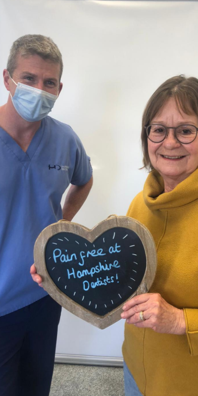 Patient with Dr Robin Hanna at Hampshire Dentists, holding a sign saying " Pain free at Hampshire Dentists" emphasising pain-free dental care.