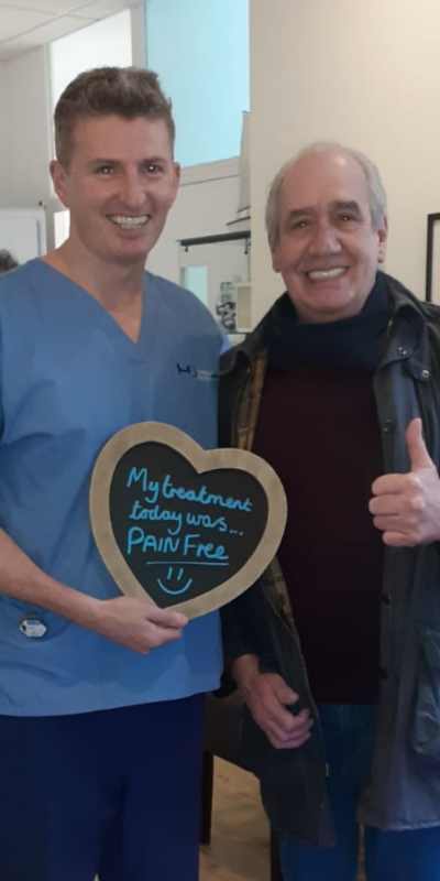 Patient with Dr Robin Hanna at Hampshire Dentists, holding a sign saying "My treatment today was pain free" emphasising pain-free dental care.