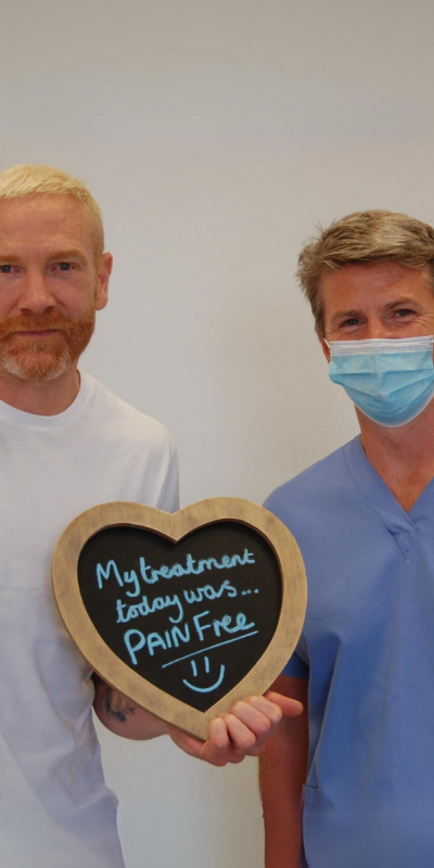 Patient with Dr Robin Hanna at Hampshire Dentists, holding a sign saying "My treatment today was pain free" emphasising pain-free dental care.
