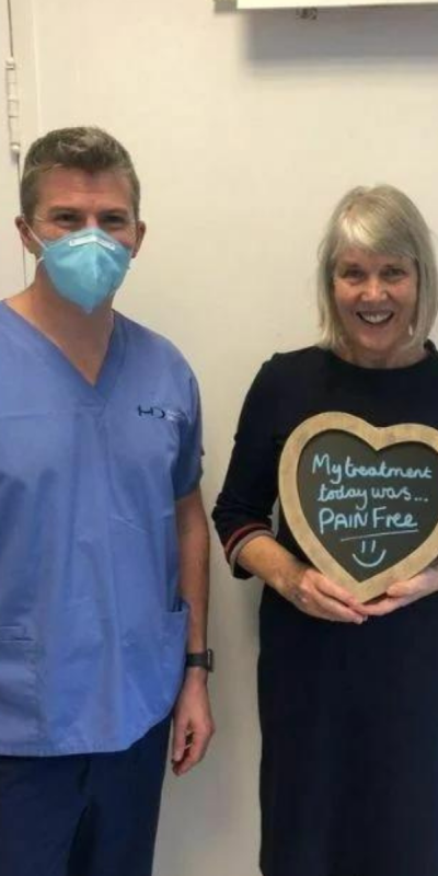 Patient with Dr Robin Hanna at Hampshire Dentists, holding a sign saying "My treatment today was pain free" emphasising pain-free dental care.