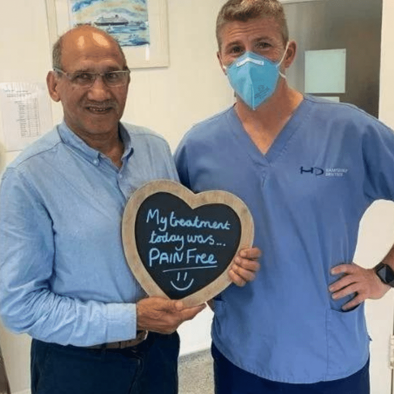 Dr Robin Hanna, Implantologist - Hampshire Dentists Pain Free Dentist in Southampton Patient with Dr Robin Hanna at Hampshire Dentists, holding a sign saying "My treatment today was pain free" emphasising pain-free dental care.