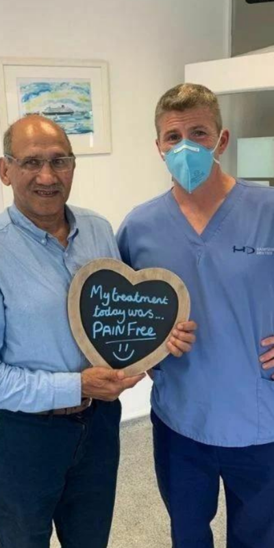 Patient with Dr Robin Hanna at Hampshire Dentists, holding a sign saying "My treatment today was pain free" emphasising pain-free dental care.