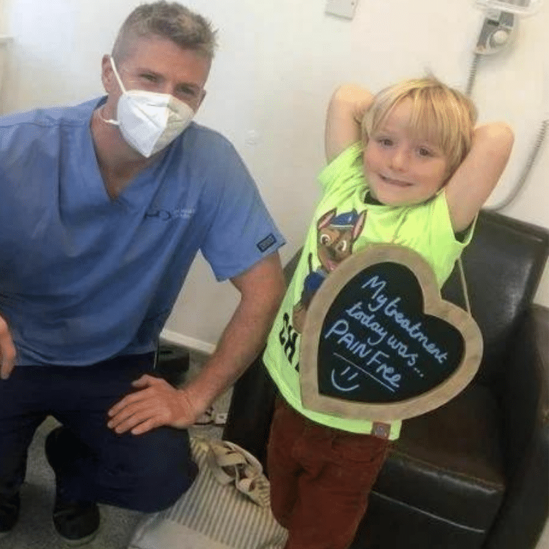 Dr Robin Hanna, Implantologist - Hampshire Dentists Pain Free Dentist in Southampton Patient with Dr Robin Hanna at Hampshire Dentists, holding a sign saying "My treatment today was pain free" emphasising pain-free dental care.