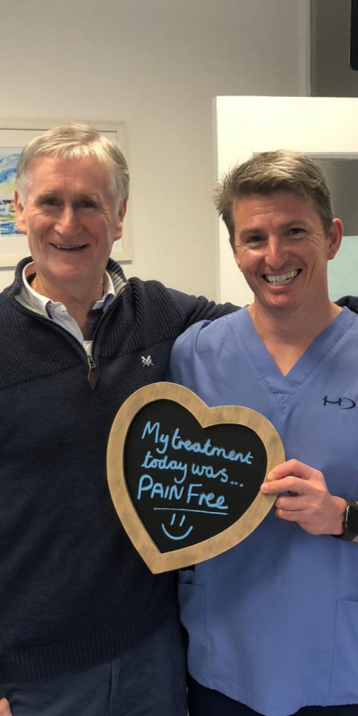 Patient with Dr Robin Hanna at Hampshire Dentists, holding a sign saying "My treatment today was pain free" emphasising pain-free dental care.