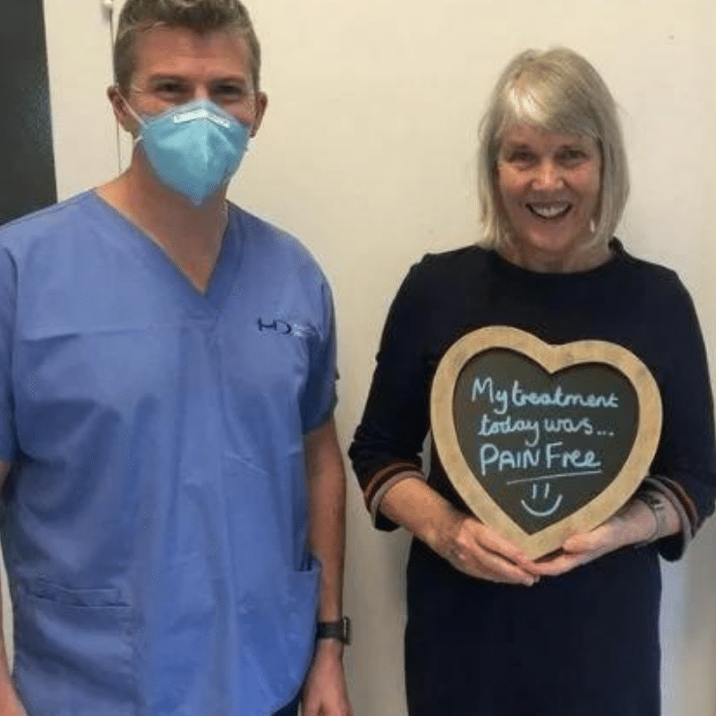 Dr Robin Hanna, Implantologist - Hampshire Dentists Pain Free Dentist in Southampton Patient with Dr Robin Hanna at Hampshire Dentists, holding a sign saying "My treatment today was pain free" emphasising pain-free dental care.