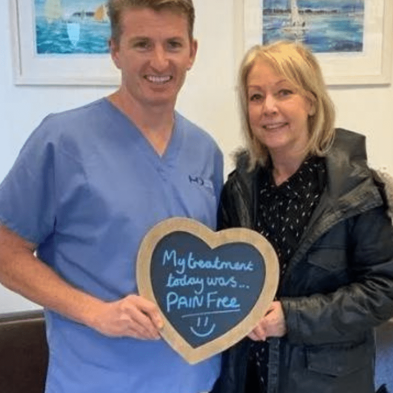 Dr Robin Hanna, Implantologist - Hampshire Dentists Pain Free Dentist in Southampton Patient with Dr Robin Hanna at Hampshire Dentists, holding a sign saying "My treatment today was pain free" emphasising pain-free dental care.