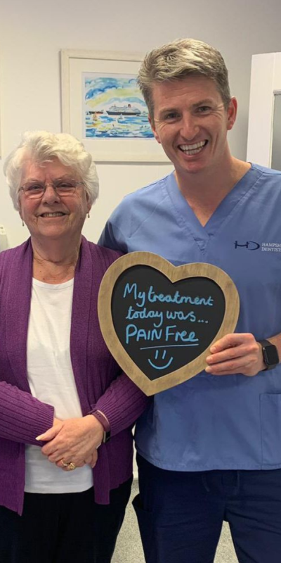 Patient with Dr Robin Hanna at Hampshire Dentists, holding a sign saying "My treatment today was pain free" emphasising pain-free dental care.