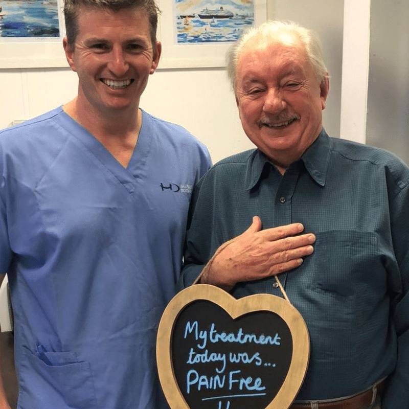 Dr Robin Hanna, Implantologist - Hampshire Dentists Pain Free Dentist in Southampton Patient with Dr Robin Hanna at Hampshire Dentists, holding a sign saying "My treatment today was pain free" emphasising pain-free dental care.