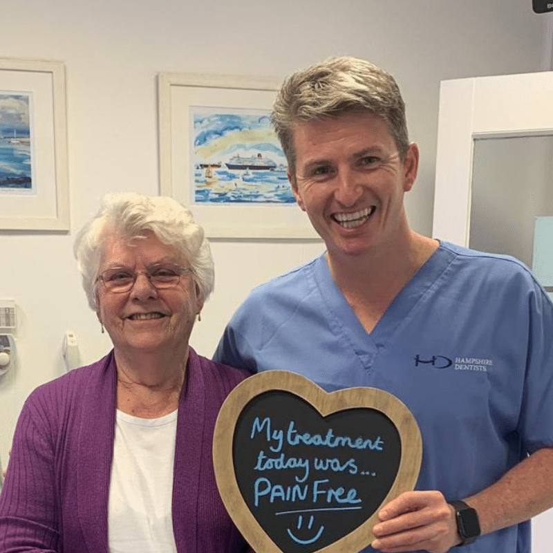 Dr Robin Hanna, Implantologist - Hampshire Dentists Pain Free Dentist in Southampton Patient with Dr Robin Hanna at Hampshire Dentists, holding a sign saying "My treatment today was pain free" emphasising pain-free dental care.