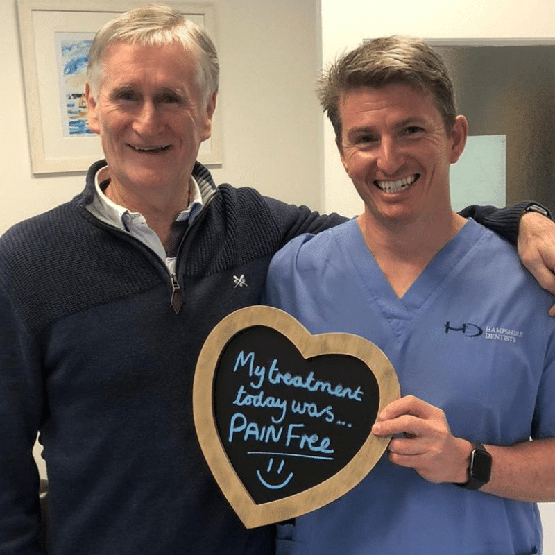 Dr Robin Hanna, Implantologist - Hampshire Dentists Pain Free Dentist in Southampton Patient with Dr Robin Hanna at Hampshire Dentists, holding a sign saying "My treatment today was pain free" emphasising pain-free dental care.